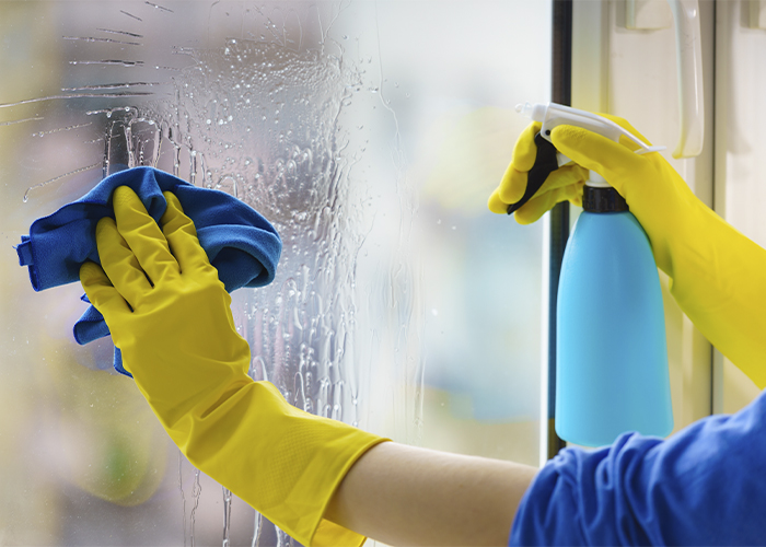 A person cleaning a window with yellow gloves, a blue cloth, and a spray bottle.